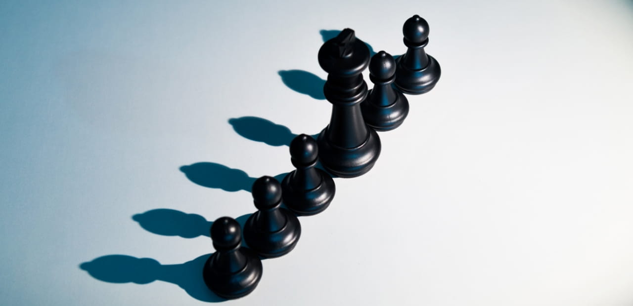 Black chess pieces arranged in a diagonal line on a light surface, casting long shadows