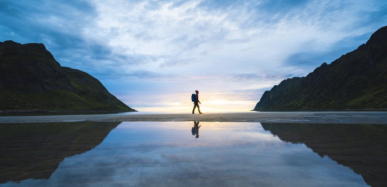 A lone person walking across a reflective shoreline between mountains at sunrise, symbolizing the six must-know principles that help new investors begin their journey with confidence and a strong foundation