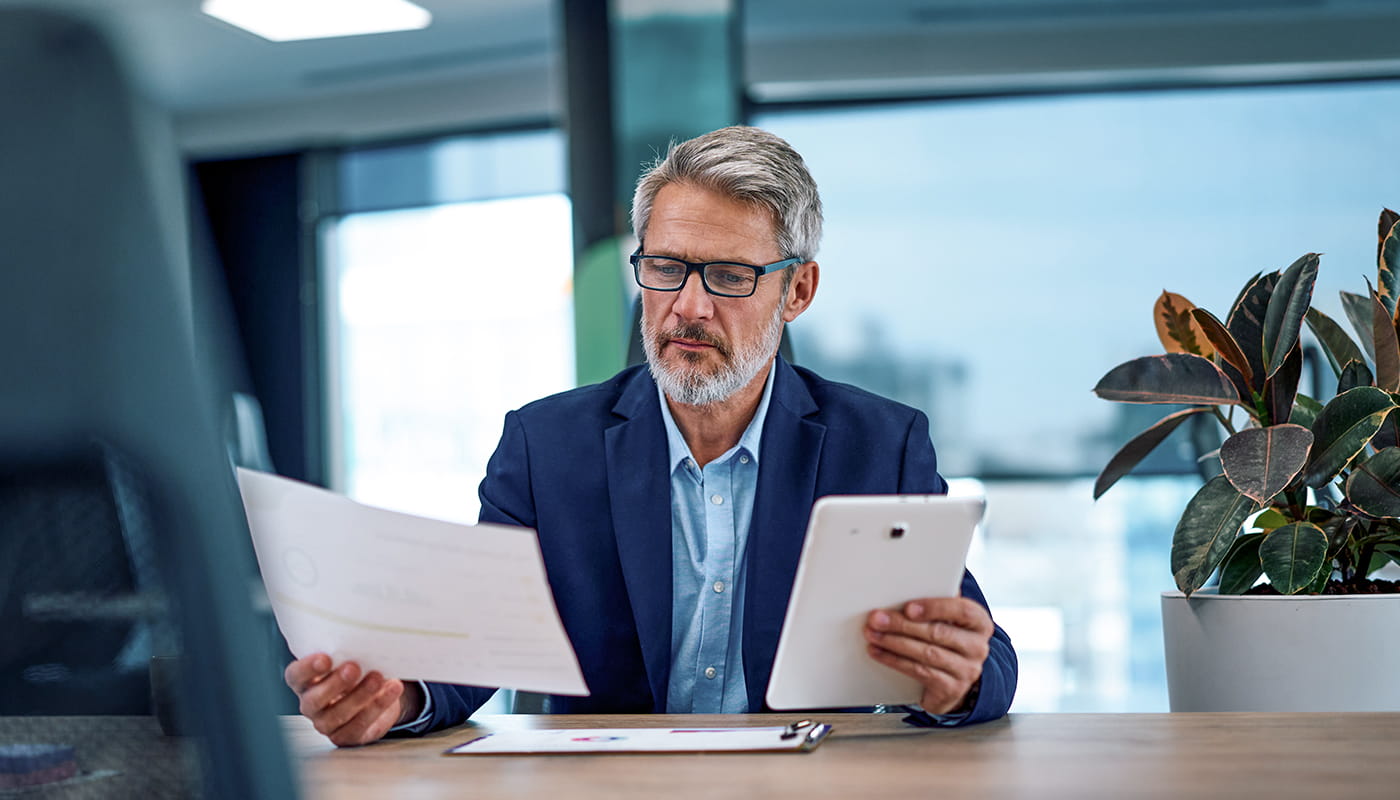 Mature adult businessman comparing documents using digital tablet in office